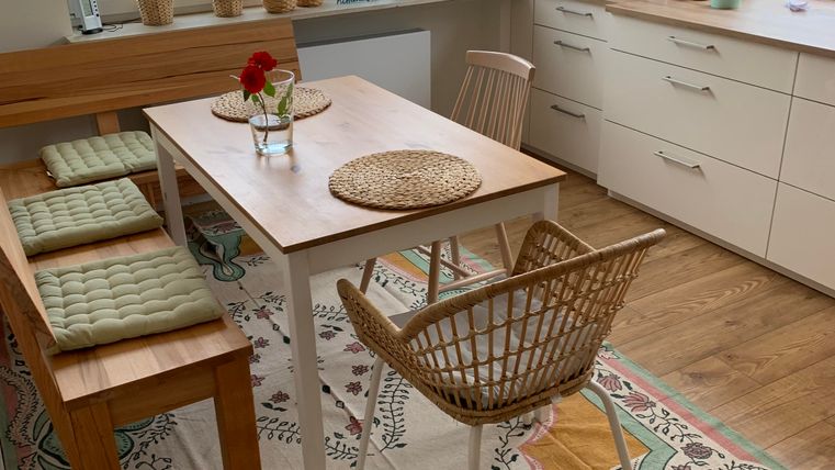 Ein gemütlicher Essbereich mit einem Holztisch und Rattan-Stühlen. Die Fenster sind hell und Pflanzen bringen Frische in den Raum.