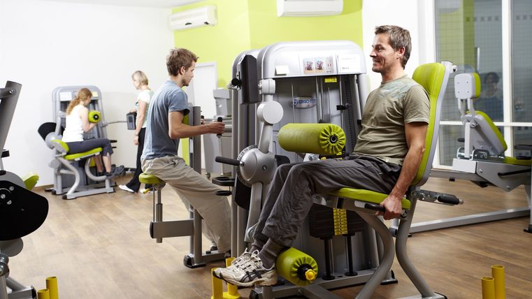Ein modernes Fitnessstudio mit mehreren Trainingsgeräten. Zwei Personen trainieren, während eine weitere Person auf einem anderen Gerät aktiv ist.