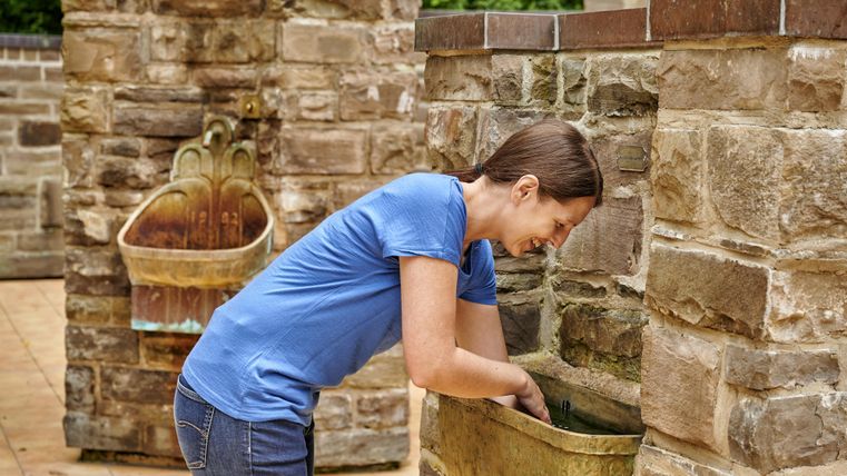 Eine Frau beugt sich über ein steinernes Waschbecken. Im Hintergrund ist eine weitere Installation sichtbar.