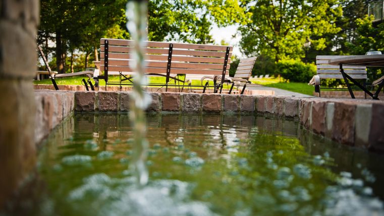 Ein ruhiger Garten mit einem kleinen Teich und fließendem Wasser. Im Hintergrund stehen Holzbänke unter großen Bäumen.