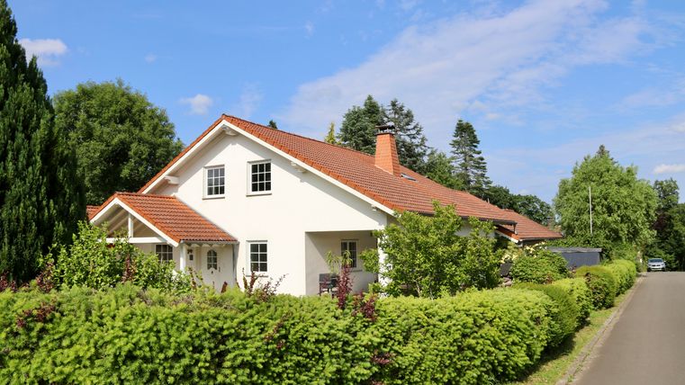 Ein schönes Einfamilienhaus mit rotem Dach und grünem Garten. Die Straße ist ruhig und von Bäumen gesäumt.