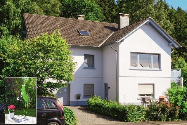 Ein modernes Haus umgeben von grüner Landschaft. Vor dem Haus steht ein kleiner Tisch mit Stühlen und Blumen.