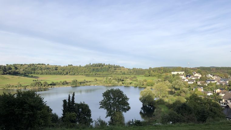 Ein ruhiger See umgeben von sanften Hügeln und Bäumen. Im Hintergrund sind einige Häuser und ein klarer Himmel zu sehen.