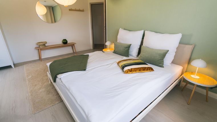 Ein modernes Schlafzimmer mit einem großen Bett und stilvollen Kissen. Die Wände sind in sanften Grüntönen gehalten, und es gibt ein gemütliches Ambiente mit minimalistischer Einrichtung.