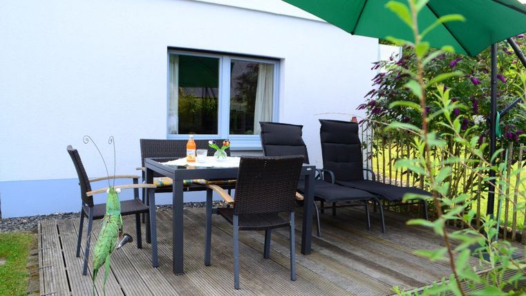 Ein gemütlicher Außenbereich mit einem Tisch und Stühlen auf einer Holzterrasse. Über dem Tisch befindet sich ein grüner Sonnenschirm und in der Nähe wachsen Pflanzen.