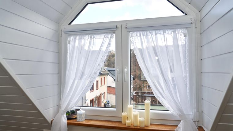 Ein Fenster mit weißen Vorhängen und einem Blick auf eine Stadtlandschaft. Auf der Fensterbank stehen einige Kerzen.
