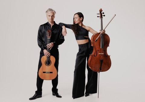 A musician with a guitar stands next to a musician with a cello. Both are wearing elegant black clothing and are posing in front of a neutral background.
