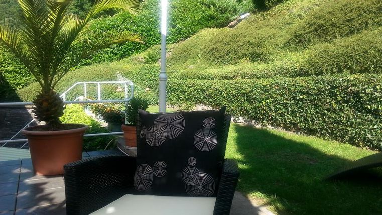 Ein gemütlicher Garten mit einem schwarzen Sessel und einem dekorativen Kissen. Im Hintergrund sind gepflegte Sträucher und eine sonnige Atmosphäre zu sehen.