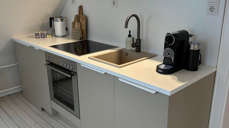 Eine moderne Küche mit hellen Schränken, einem Edelstahlherd und einem Waschbecken. Auf der Arbeitsplatte stehen eine Kaffeemaschine und dekorative Elemente.