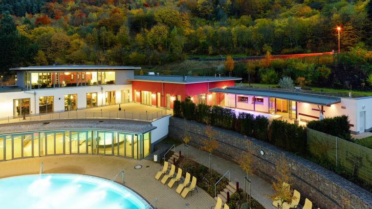 Ein modernes Wellnessresort mit einem Pool und Liegen im Vordergrund. Im Hintergrund sind die bunten Bäume der Landschaft zu sehen.