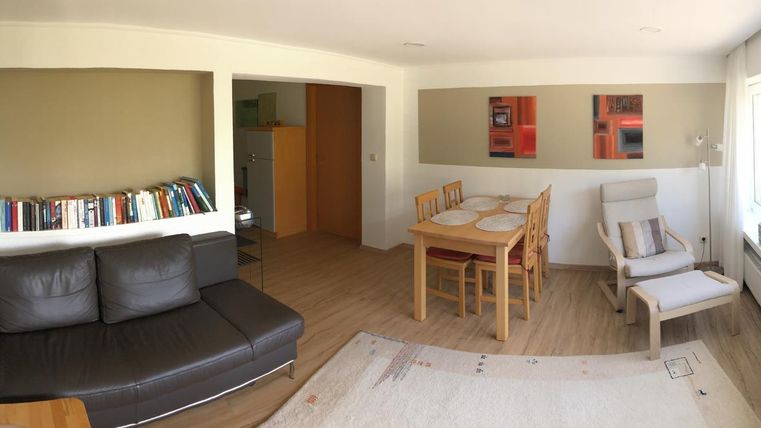 Ein helles und einladendes Wohnzimmer mit einem schwarzen Sofa, einem Esstisch und modernen Kunstwerken an der Wand. Große Fenster sorgen für viel Tageslicht und eine angenehme Atmosphäre.