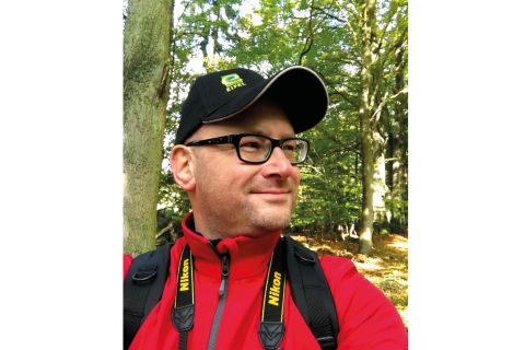Ein Mann mit einer Kamera steht in einem Wald. Er trägt eine rote Jacke und eine schwarze Mütze.