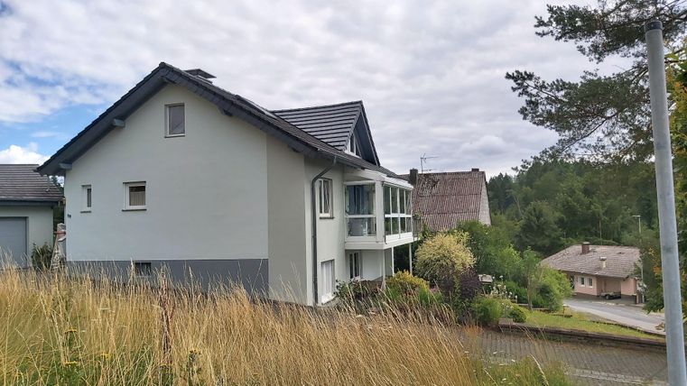 Ein modernes Haus mit zwei Stockwerken und einem Balkon. Im Hintergrund sind Bäume und andere Gebäude zu sehen.