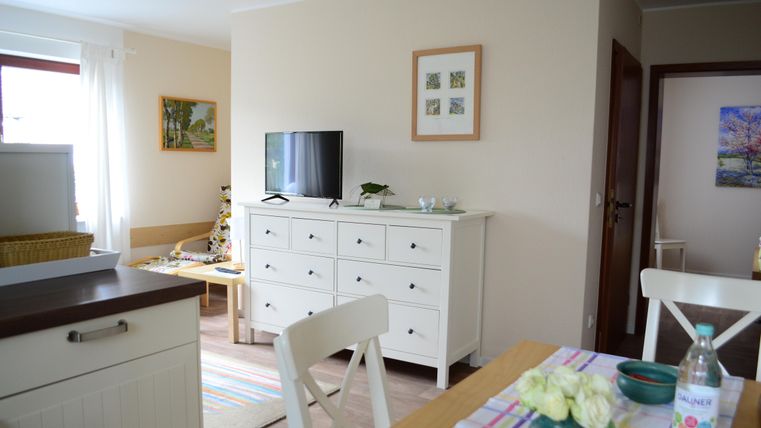 Ein helles, modernes Wohnzimmer mit einem Fernseher und einer weißen Kommode. Der Raum hat einen einladenden Esstisch mit Blumen und einem Wasserkrug.
