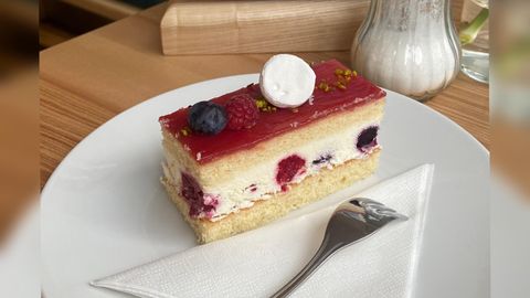 Ein Stück Sahnetorte mit einer fruchtigen Schicht aus Himbeeren und Heidelbeeren. Der Kuchen liegt auf einem weißen Teller mit einem Löffel und einer Serviette.