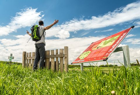 Ein Wanderer steht mit ausgebreiteten Armen vor einem Holzzaun auf dem Vulcano-Pfad. Ein Informationsschild ist im Vordergrund sichtbar.
