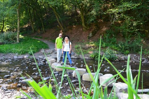 Ein Paar steht auf Steinen in einem kleinen Bach. Die Umgebung ist grün und bewaldet, ideal für einen entspannten Ausflug in die Natur.