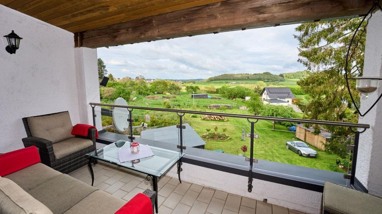 Eine gemütliche Terrasse mit einem Glas Tisch und bequemen Möbeln. Der Blick geht auf eine grüne Landschaft mit Gärten und Hügeln im Hintergrund.