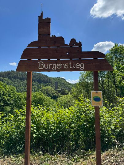 Ein Schild des Burgenstiegs, das eine Burgruine darstellt, steht vor einer bewaldeten Landschaft. Der Himmel ist blau mit einigen Wolken.
