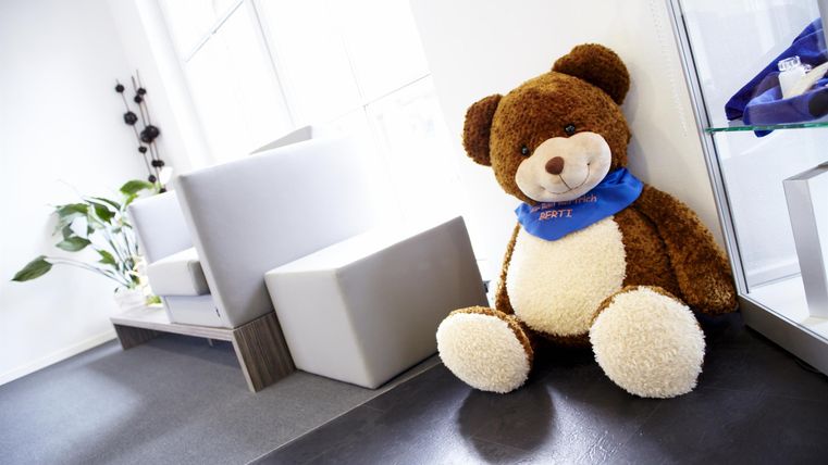 Ein großer, weicher Teddybär sitzt in einem hellen Raum. Daneben gibt es moderne Möbel und ein paar Pflanzen.