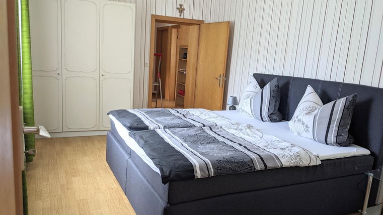 Ein modernes Schlafzimmer mit einem großen Bett und hellen Holzfußböden. An der Wand sind helle Paneele und ein Kleiderschrank sichtbar.