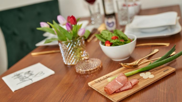 Ein schön gedeckter Tisch mit frischen Zutaten. Darauf sind Teller, eine Salatschüssel, eine Blumenvase und fein angerichteter Schinken zu sehen.