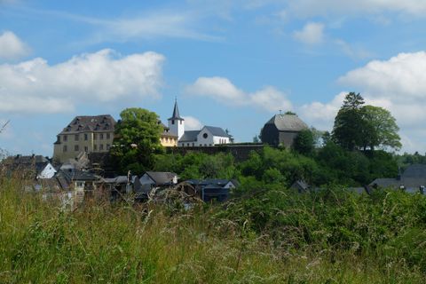 Ein malerisches Dorf auf einem Hügel mit historischen Gebäuden und einem Kirchturm. Umgeben von grünen Bäumen und einem klaren Himmel.
