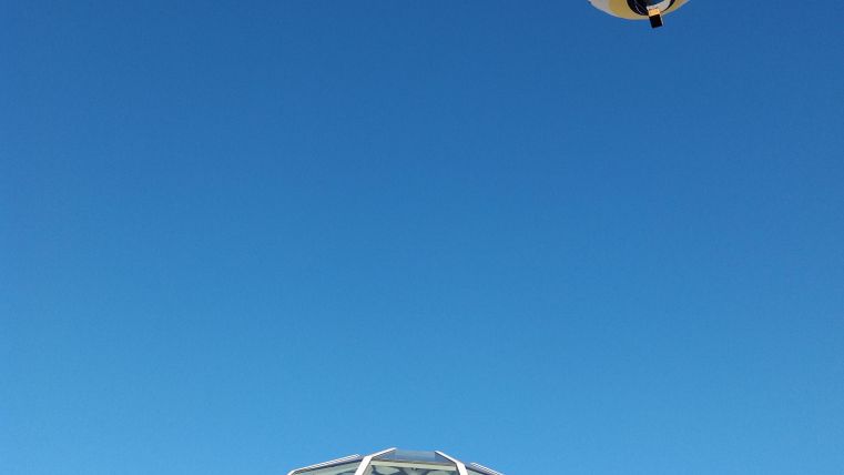 A hot air balloon floats high in the sky above a modern building. The clear blue sky provides a beautiful backdrop.