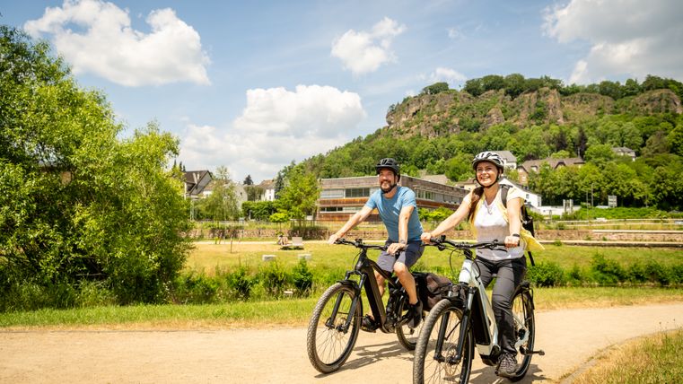 Two people ride bicycles on a path surrounded by green countryside and hills.