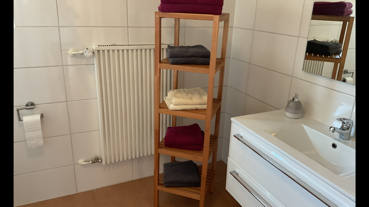 Ein modernes Badezimmer mit einem Holzregal, das ordentlich gefaltete Handtücher in verschiedenen Farben zeigt. Im Hintergrund sind ein Waschbecken und ein Spiegel zu sehen.
