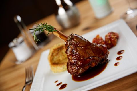 Ein zartes Lammkeulensteak auf einem weißen Teller, garniert mit Rosmarin. Daneben sind einige Kirschtomaten und ein Stück Polenta.