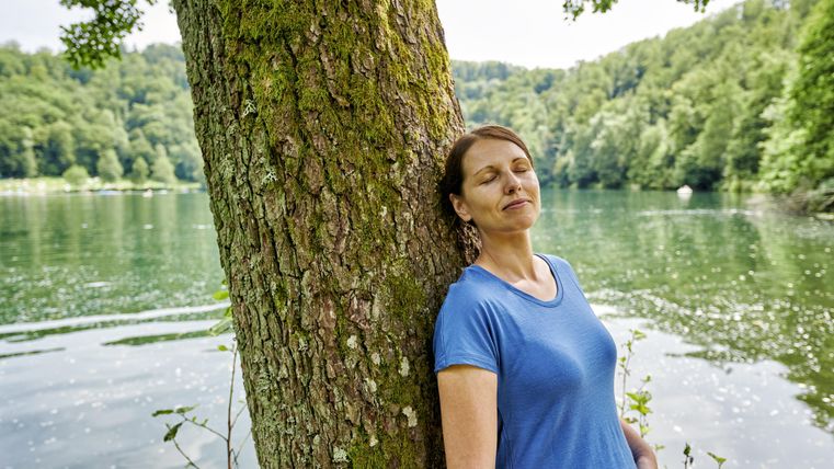 Eine Frau lehnt entspannt an einem Baum am Ufer eines ruhigen Sees. Die Umgebung ist grün und friedlich.