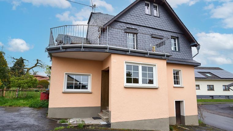Ein modernes Haus mit einem hellen, verputzten Erdgeschoss und einem schiefergedeckten Obergeschoss. Das Gebäude hat einen Balkon und befindet sich in einer ruhigen Umgebung.