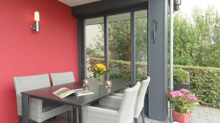 Ein stilvoller Balkon mit einem Tisch und Stühlen aus Rattan. Auf dem Tisch stehen Blumen und Kerzen, während im Hintergrund grünes Laub sichtbar ist.