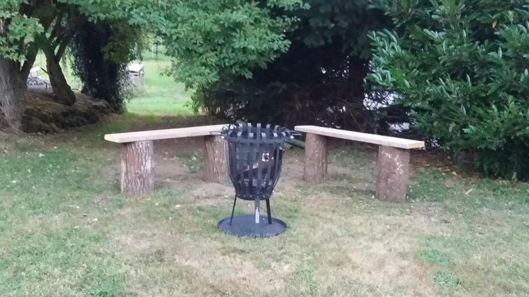 Eine gemütliche Sitzecke im Freien mit zwei Holzbänken und einem Feuerkorb in der Mitte. Umgeben von Grünflächen und Bäumen.