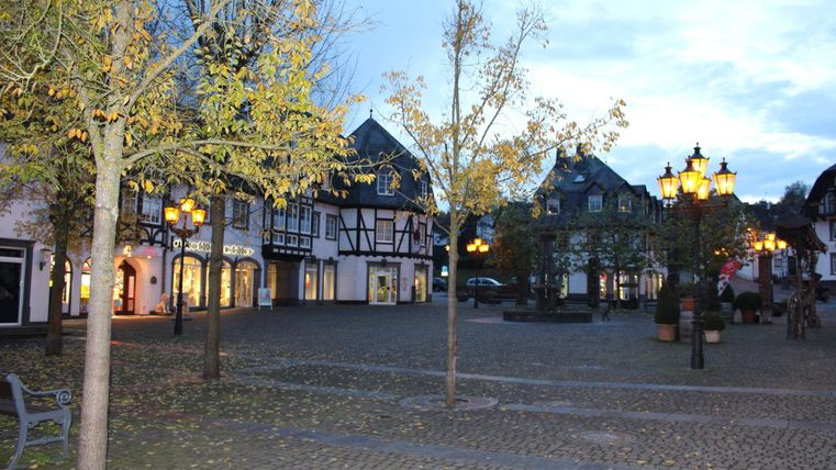 Ein malerischer Platz mit traditionellen Fachwerkhäusern und herbstlichen Bäumen. Am Abend leuchten die Straßenlaternen und schaffen eine einladende Atmosphäre.