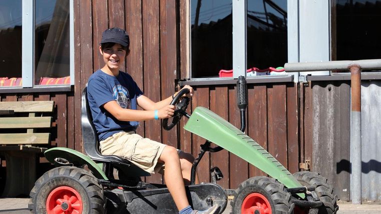 Ein Junge sitzt auf einem kleinen Traktor und lächelt. Im Hintergrund sind Holzpaneele und Fenster zu sehen.