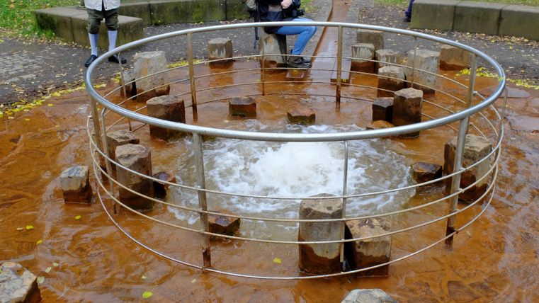 Ein runder Wasserbereich umgeben von Holzstämmen. In der Mitte sprudelt das Wasser.