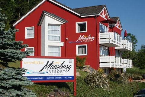 Ein rotes Gebäude mit dem Namen "Mahnberg Resort" und mehreren Balkonen. Ein Willkommensschild steht im Vordergrund, umgeben von grüner Landschaft.