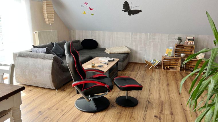 Ein gemütlicher Wohnraum mit einem grauen Sofa, einem modernen Stuhl und einem Tisch. An der Wand sind dekorative Schmetterlinge und Pflanzen zu sehen.