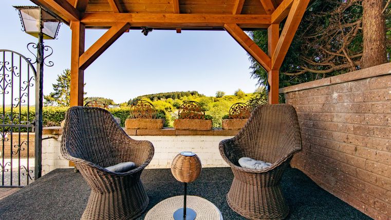 Eine gemütliche Terrasse mit zwei geflochtenen Stühlen und einem kleinen Tisch. Im Hintergrund öffnet sich eine grüne Landschaft unter einem klaren Himmel.