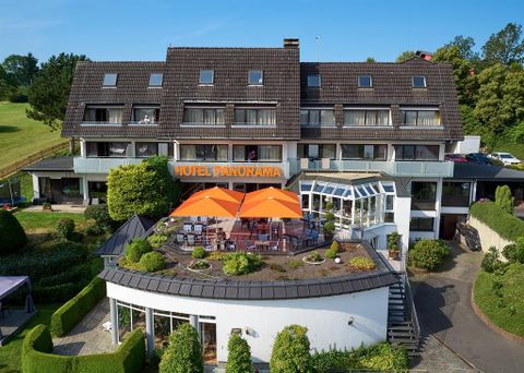 Ein modernes Hotel mit dem Namen „Hotel Panorama“ und einer einladenden Terrasse. Die Terrasse ist mit orangefarbenen Sonnenschirmen und grünen Pflanzen dekoriert.