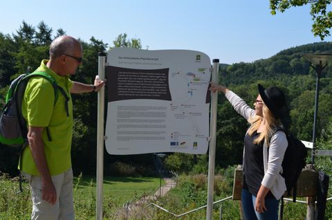 Zwei Wanderer betrachten ein Informationstafel in der Natur. Im Hintergrund sind Bäume und Hügel zu sehen.