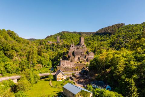 Eine beeindruckende Burgruine umgeben von grünen Wäldern. Im Vordergrund sind einige Häuser und eine Straße zu sehen.