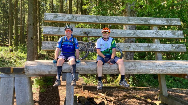 Zwei Radfahrer sitzen auf einer großen Holzbank im Wald. Im Hintergrund sind Bäume zu sehen, und die Sonne scheint.