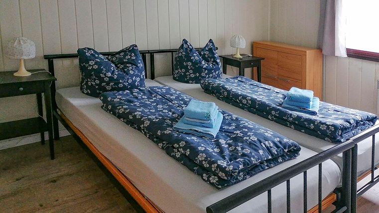 Ein gemütliches Zimmer mit zwei Betten, jeweils mit blauen Bettdecken und Kissen. Neben den Betten liegen frische Handtücher und es gibt kleine Nachttische mit Lampen.