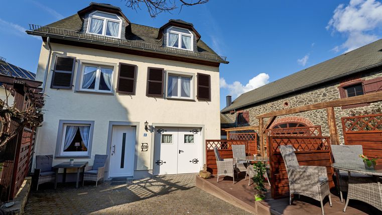 Ein gemütliches Haus mit einem klaren blauen Himmel im Hintergrund. Vor dem Haus steht eine Terrasse mit Gartenmöbeln.