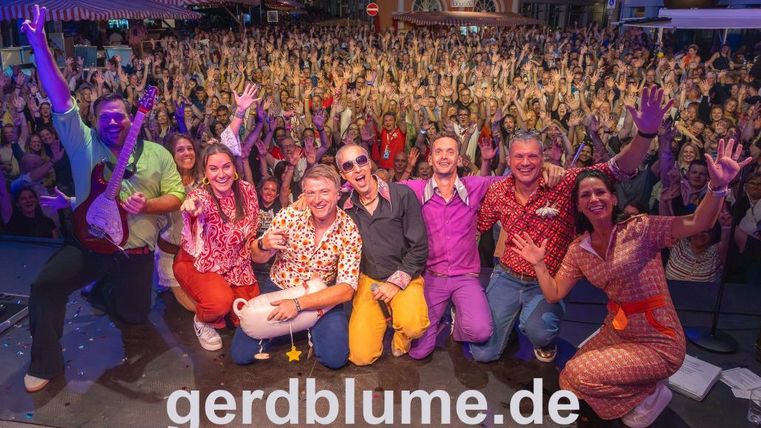A cheerful group of people stands on a stage and poses for the photo. In the background, a large crowd of spectators can be seen, enthusiastically applauding.