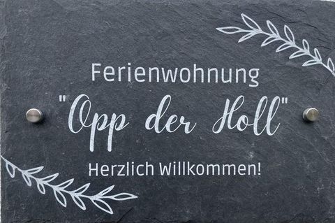 Ein Schild für eine Ferienwohnung mit dem Namen "Opp der Holl". Es heißt "Herzlich Willkommen!" und hat eine dekorative Schrift und Pflanzenmotive.
