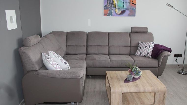 Ein modernes Wohnzimmer mit einer grauen Couch und einem Holztisch. An der Wand hängt ein buntes Gemälde und es gibt dekorative Kissen auf der Couch.